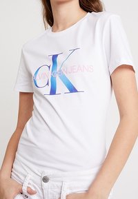 Biała bawełniana koszulka z niebieskim gradientowym logo "CK" i różowym napisem "CALVIN KLEIN JEANS". Stylizowana z krótkimi rękawami i dopasowanym krojem.