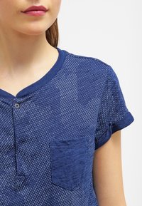 Námořnická modrá krátkosrstá henley košile s texturovanou tkaninou, s malými knoflíky a kapsou. Měkký, lehký materiál s vzorovaným designem.