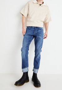 Beige kortärmad polo med vit krage, i kombination med blå denimjeans och svarta ankelstövlar med gula sömmar. Jeansen är uppvikta.