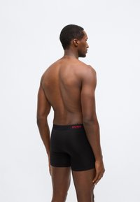 Melni bokserveida šorti ar ciešu piegulumu, uz vidukļa lentes redzams sarkans logotips. Gluda auduma un vienkārša dizaina, skatīti no mugurpuses.