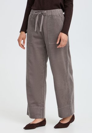 Femme portant un pantalon large en velours côtelé gris avec taille à cordon et grandes poches avant, associé à des ballerines marron foncé et un haut.
