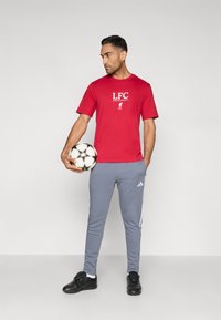 Κόκκινο κοντομάνικο μπλουζάκι με το λογότυπο "LFC", σε συνδυασμό με γκρι φόρμες με λευκές πλευρικές ρίγες. Μαύρα αθλητικά παπούτσια. Κρατά μια μπάλα ποδοσφαίρου.