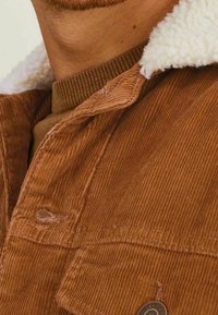 Braune Kordjacke mit einem weichen, weißen Fleece-Kragen. Merkmale sind sichtbare Nähte und ein abgerundetes Metallelement nahe dem Saum.