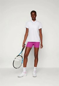 Hvit polo-skjorte og rosa trening-shorts. Modellen holder en tenniskølle. Klærne er laget av pustende stoff, egnet for sportsaktiviteter.