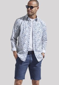 Weißes Hemd mit blauem abstraktem Muster, über einem weißen T-Shirt getragen, kombiniert mit dunkelblauen Shorts. Accessoires mit dunklen Sonnenbrillen.