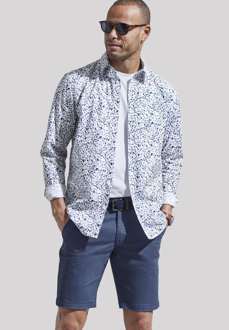 Weißes Hemd mit blauem abstraktem Muster, über einem weißen T-Shirt getragen, kombiniert mit dunkelblauen Shorts. Accessoires mit dunklen Sonnenbrillen.