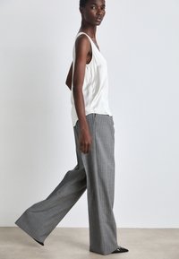 Satin valkoinen hihaton paita yhdistettynä harmaisiin raidoitettuihin leveälahkeisiin housuihin, joissa on sileä pinta ja rento istuvuus, täydentävät mustat kärkitossut.