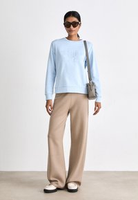 Világoskék, kereknyakú pulóver, dombornyomott logóval, bézs bő szárú nadrággal és fehér, fekete talpú cipővel párosítva. Kiegészítő: kis szürke kézitáska.