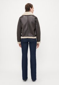 Kvinne med kort hår som har på seg en mørkebrun lammeskinnsjakke og mørkeblå jeans står med ryggen til mot en ensfarget bakgrunn.