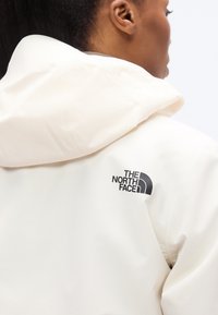 Λευκό αδιάβροχο μπουφάν με κουκούλα, με μαύρο λογότυπο στον αριστερό ώμο. Λείο ύφασμα, κομψός σχεδιασμός και μινιμαλιστικό στυλ.