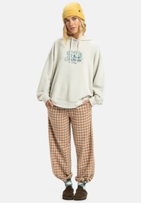 Overmaat crèmekleurige hoodie met een zak aan de voorkant, featuring een bloemmotief. Bruin en wit geblokte relaxte broek met elastische boorden. Bruine slippers.
