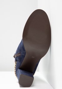 Chaussure à talons hauts en daim bleu marine avec une semelle brune texturée, fermeture éclair sur le côté et accent en chaîne dorée près du talon. Taille indiquée comme 37.