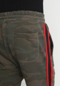 Kalhoty s camouflage vzorem, elastickým pasem a zadní kapsou, se svislými černými a červenými pruhy po stranách na jednoduchém pozadí.