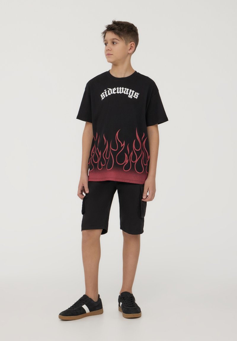 T-shirt nera con stampa di fiamme rosse e testo bianco "di lato", abbinata a pantaloni cargo neri e sneaker nere con suole marroni.