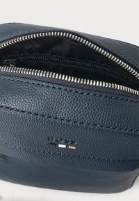 Tengerészkék bőr tasak texturált felülettel, ezüst cipzárral, fekete szövet béléssel és finom fekete, fehér és narancssárga márkajelzéssel.