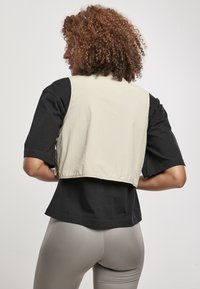 Beżowa krótka kamizelka na czarną koszulę z krótkimi rękawami; zestawiona z błyszczącymi, jasnoszarymi legginsami. Tekstura dotykowa i swobodny design.