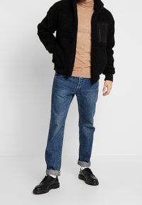 Sort fleecejakke med lynlås-lomme, beige t-shirt, blå denimjeans og sorte snøresko. Jakken har ribbede manchetter og kant.