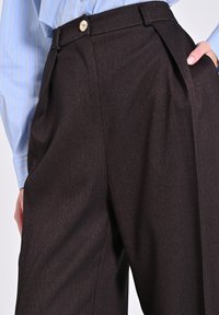 Braune Hosen im Fischgrat-Muster mit einem gefalteten Design, einem goldenen Knopfverschluss und glatter Textur. Kombiniert mit einem hellblauen gestreiften Hemd.