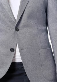 Blazer grigio con texture, due bottoni neri, rever a punta e tasca frontale singola. Vestibilità su misura con fodera liscia.