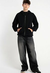 Czarna bluza z zamkiem i fakturowym wykończeniem, zestawiona z luźnymi, szarymi dżinsami o wyblakłym wzorze. Całość dopełniają czarne sneakersy.