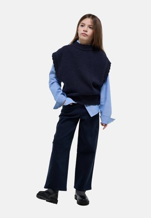 Jeune fille portant un gilet en maille bleu marine sur une chemise bleu clair, un pantalon large foncé et des chaussures noires, debout avec une main dans la poche.