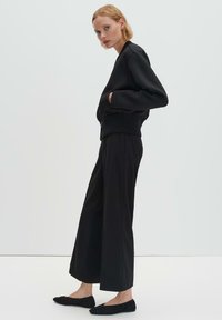 Blouson aviateur noir court avec ourlet côtelé, associé à un pantalon large noir ample et à des chaussures noires à enfiler. Texture de tissu lisse.