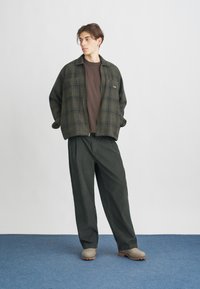 Zeleno karo zip-jakna čez rjavo srajco, v kombinaciji z ohlapnimi temno zelenimi hlačami in svetlo sivimi klobuki na modrem preprogem tlaku.