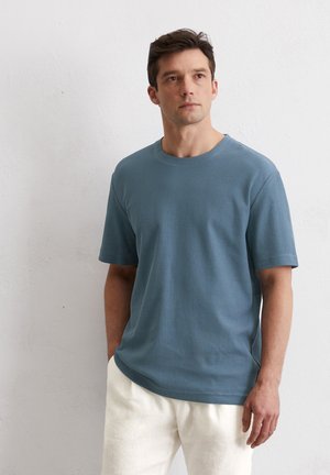 Helles blaues kurzärmeliges T-Shirt aus strukturiertem Stoff, mit Rundhalsausschnitt und entspannter Passform, kombiniert mit hellen Hosen.