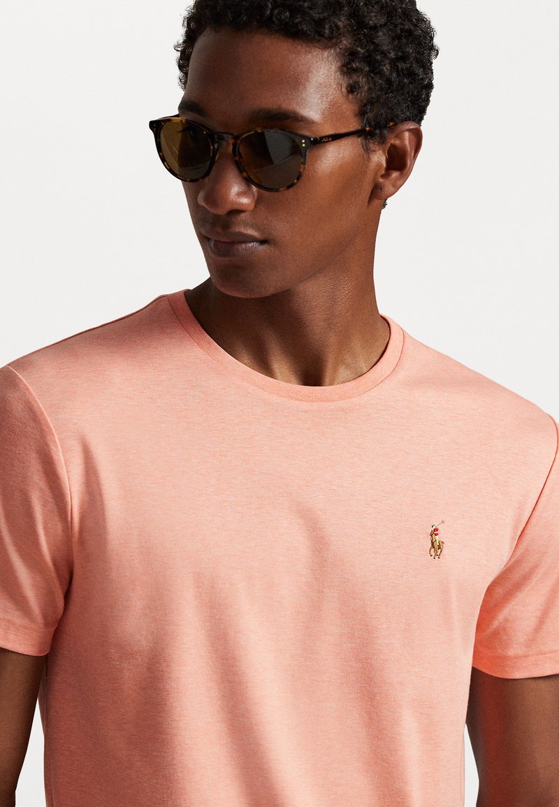 T-shirt en coton rose clair avec un col rond et un petit logo brodé sur la poitrine gauche. Portée avec des lunettes de soleil en écaille.