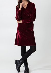 Burgundské sametové zavinovací šaty s dlouhými rukávy, ke kolenům, s nařasenými detaily na ramenou. V páru s černými kotníkovými botami.