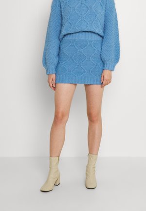 Kvinde iført en blå strikket sweater og matchende mininederdel med beige ankelstøvler, stående foran en ensfarvet hvid baggrund.