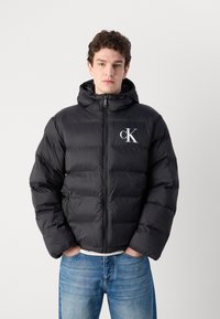 Schwarze Steppjacke mit Kapuze, ausgestattet mit einem Reißverschluss vorne und elastischen Bündchen. Enthält ein weißes "cK"-Logo auf der linken Brust. Texturiertes, gestepptes Material.