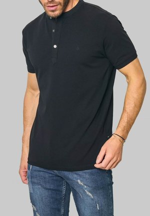 T-shirt henley à manches courtes en coton noir avec un plastron à trois boutons, ourlet arrondi et logo subtilement détaillé. Porté avec un jean bleu déchiré.