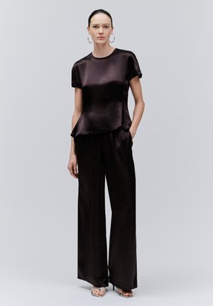 Femeie în picioare, purtând un top scurt cu mâneci din satin negru și pantaloni evazați, cu cercei argintii cerc și pantofi cu toc deschis argintii.