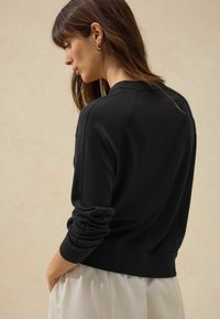 Pull noir à coupe décontractée, avec des poignets et un ourlet côtelés. Présente des manches raglan et un tissu doux et lisse. Porté par-dessus un pantalon de couleur claire.