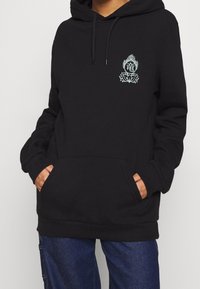 Czarna bluza z kapturem z przednią kieszenią, regulowanym kapturem oraz białym wzorem graficznym z elementami roślinnymi i symbolami na klatce piersiowej.