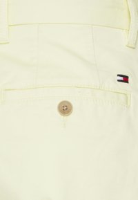 Poche arrière de pantalon jaune clair avec un bouton marron et un petit logo Tommy Hilfiger rouge, blanc et bleu sur le côté droit.