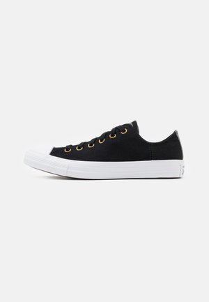 Black canvas sneaker with white rubber sole, featuring gold eyelets and round laces. Low-top design with textured sides and classic shape.