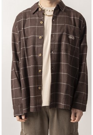 Hombre con camisa marrón a cuadros desabrochada sobre camiseta blanca, cadena de oro y collar de perlas, pantalones cargo marrones visibles.