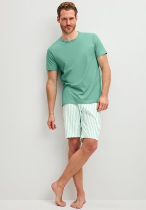 Mann steht barfuß vor einer weißen Wand und trägt ein grünes kurzärmeliges Hemd sowie weiße Shorts mit dünnen grünen vertikalen Streifen.