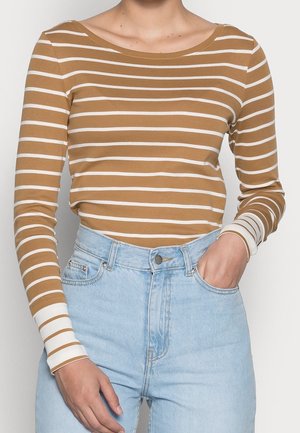 Vrouw met een bruin-wit horizontaal gestreept shirt met lange mouwen, ingestopt in een hooggetailleerde lichtblauwe spijkerbroek, met één hand in de zak.