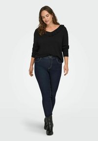 Žena u crnom dugom rukavu s naboranim izrezom, tamnim uskim trapericama i crnim čizmama do gležnja, stoji naslonjena na jednostavnu pozadinu.