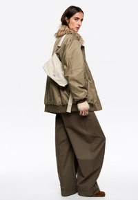 Oliwkowa kurtka bomberka z futrzanym wykończeniem, zestawiona z szerokimi khaki spodniami. Mała kremowa torebka wisi na jednym ramieniu. Srebrne pierścionki zdobią palce.