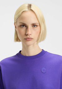 Purple sweatshirt with a round, tonal smiley logo. Features a crew neck and smooth fabric texture. Close-up, minimal background.
