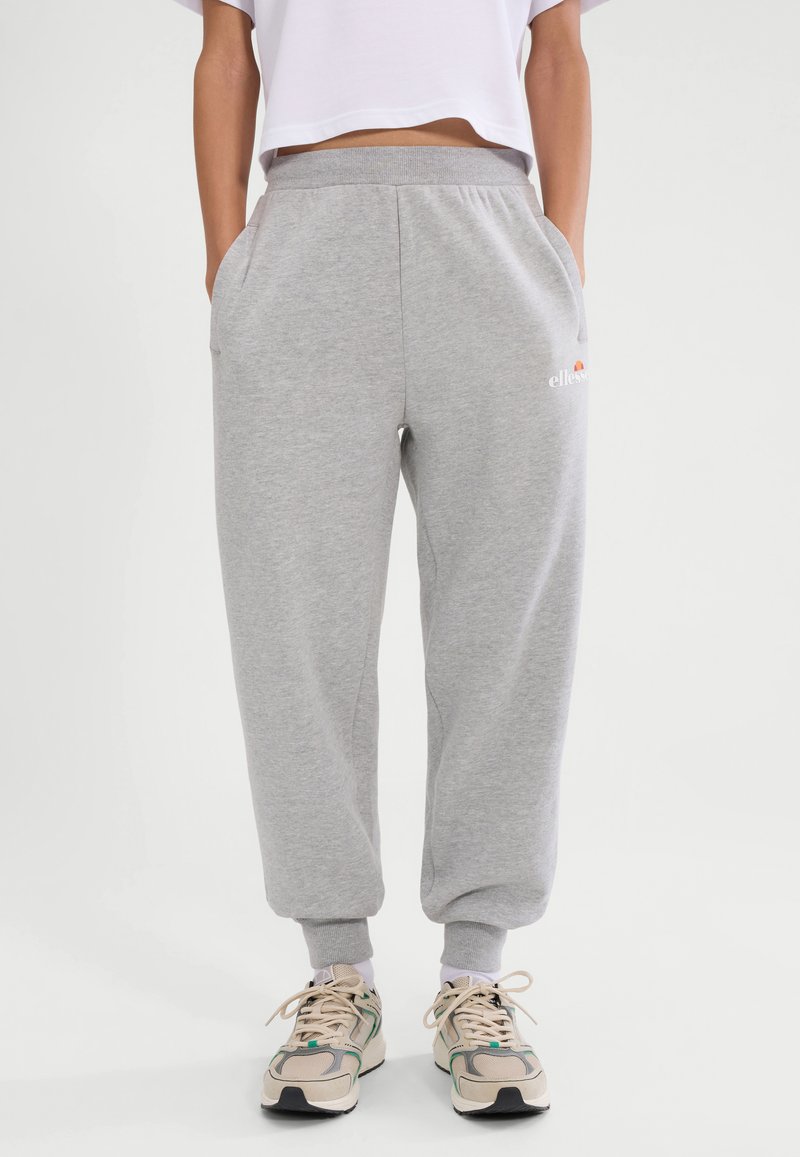 Osoba ubrana w jasnoszare spodnie dresowe z logo na udzie, białą krótka koszulkę oraz beżowe trampki ze sznurowadłami w białym kolorze, stojąca z rękami w kieszeniach.