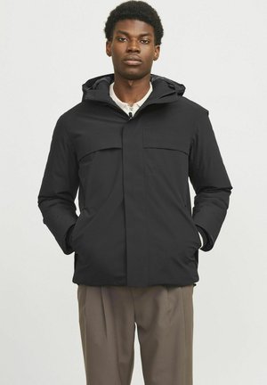 Chaqueta negra con capucha, hecha de un tejido suave y ligero. Presenta bolsillos frontales y un diseño recto. Llevada sobre pantalones beiges.