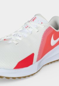 Λευκό και κόκκινο αθλητικό παπούτσι Nike με κορδόνια, υφή επάνω μέρος και καφέ λαστιχένια σόλα, εμφανίζεται από πλευρική γωνία μπροστά.