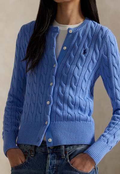 Femeie purtând un pulover cu nasturi bleu, tricotat cu model în relief, peste un top alb și blugi albaștri, cu mâinile în buzunare, pe un fundal neutru.