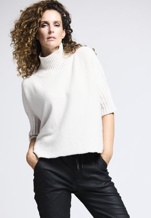 Gestrickter cremfarbener Pullover mit einem hohen Rippkragen und kurzen Ärmeln, kombiniert mit schwarzen, enganliegenden Hosen, die eine elegante Silhouette schaffen.