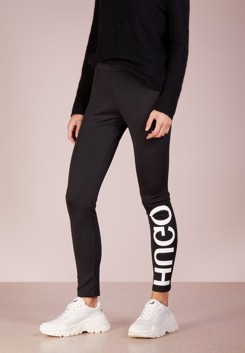 Mustat leggingsit, joissa on vasemmassa jalassa valkoinen "Hugo" -logo, yhdistettynä mustaan, teksturoituun pitkähihaiseen neuleeseen ja valkoisiin paksupohjaisiin tennareihin.